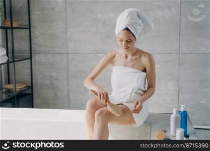 Young woman wrapped in towel massages hip with natural massaging brush, sitting on the side of bathtub in modern bathroom after the shower. Cellulite treatment, skincare, body care concept.. Woman wrapped in towel massages hip with massaging brush in bathroom. Cellulite treatment, body care