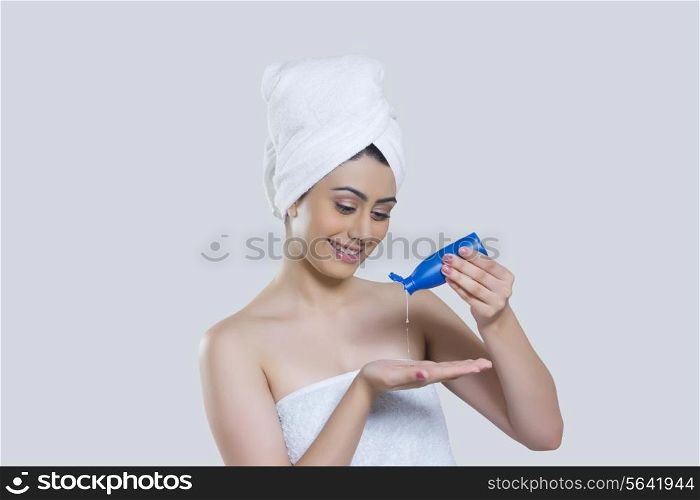 Young woman wrapped in towel pouring hair oil on palm over gray background