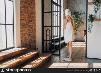 Young woman wrapped in towel taking step onto electronic floor scale, measuring weight after shower at home on sunny day. Girl checking weight loss. Slimming and healthy lifestyle concept.. Girl wrapped in towel measuring weight using electronic floor scale after shower. Slimming, wellness