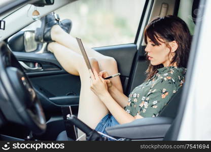 Young woman writing in a notebook with a pen in a white car pulling her feet out the window. Female wearing flowered shirt and shorts.. Woman writing in a notebook with a pen in a white car