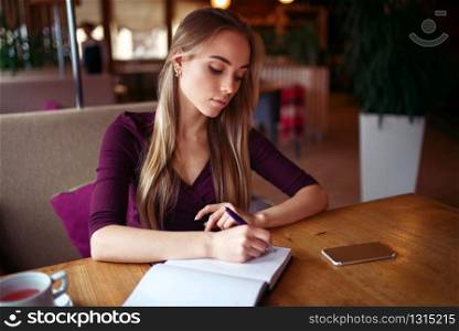 Young woman writing in her notebook, mobile phone on the table, cafe on background
