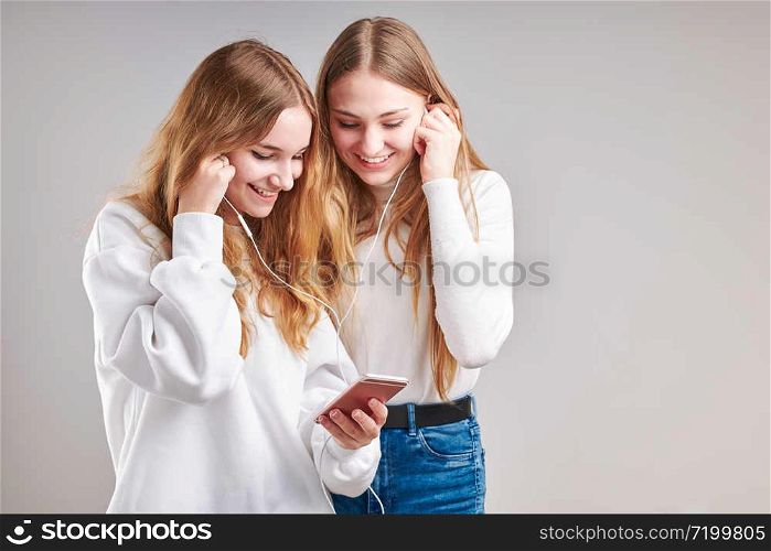 Young women girls listening to music together streaming content having fun watching video enjoying video chat talking with friends making gestures faces using smartphone earphones headphones standing over plain grey background