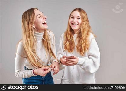 Young women girls listening to music together streaming content having fun watching video enjoying video chat talking with friends making gestures faces using smartphone earphones headphones standing over plain grey background