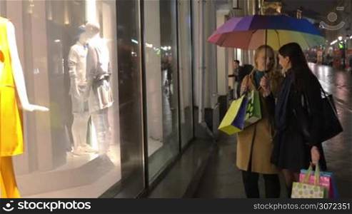Young women having shopping evening. They holding umbrella and shopping bags while looking at show-window and discussing clothing presented on mannequins