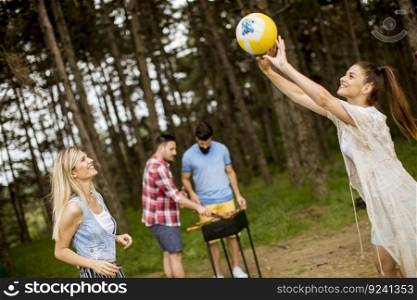 Young women playing volleyball on picnik in spring nature while men preparing grill