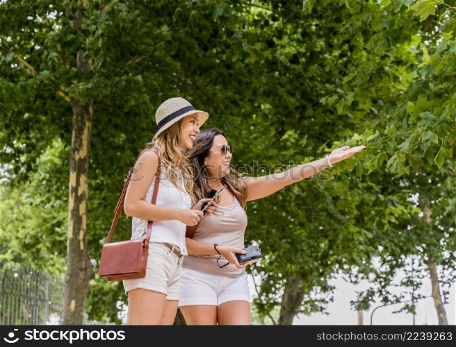 young women wearing hat carrying shoulder bag looking her female friend showing something