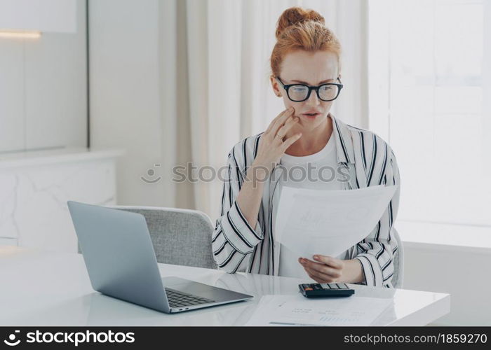 Young worried depressed woman with having financial problems, sitting at table with laptop and calculator while managing monthly budget, frustrated redhead female reading letter with bank notification. Young depressed woman with having financial problems, sitting at table with laptop and calculator