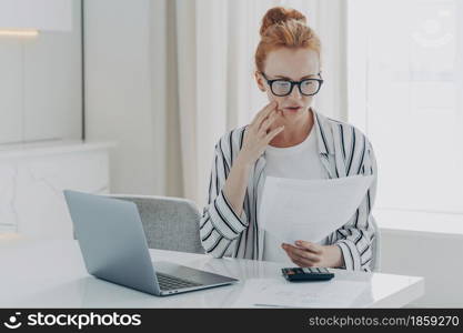 Young worried depressed woman with having financial problems, sitting at table with laptop and calculator while managing monthly budget, frustrated redhead female reading letter with bank notification. Young depressed woman with having financial problems, sitting at table with laptop and calculator