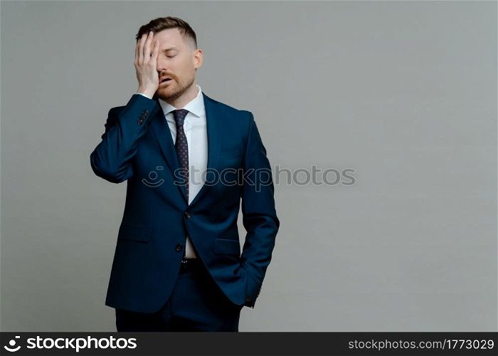 Young worried man manager in formal wear feeling frustrated and upset about business failure, holding hand on face and keeping eyes closed while standing against grey background, copy space for text. Upset man in suit feeling depressed of business failure