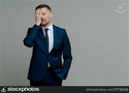 Young worried man manager in formal wear feeling frustrated and upset about business failure, holding hand on face and keeping eyes closed while standing against grey background, copy space for text. Upset man in suit feeling depressed of business failure