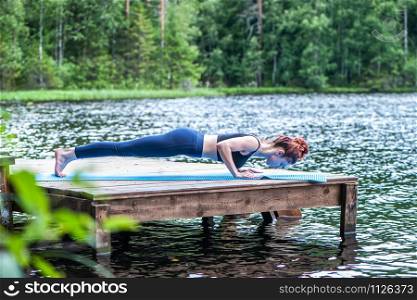 Young yogi girl practicing yoga, doing four limbed staff, chaturanga dandasana pose, on the lake. Concept of healthy life and natural balance