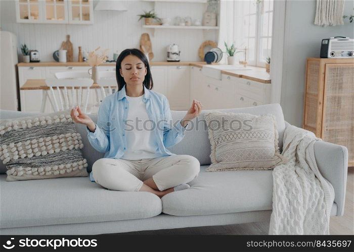 Zen, tranquility and breath control. Spanish woman is practicing yoga gymnastics in living room. Girl sitting on couch in lotus pose and meditating with her eyes closed. Relaxation and stress relief.. Zen, tranquility and breath control. Spanish woman is practicing yoga gymnastics in living room.