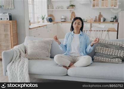 Zen, tranquility and breath control. Spanish woman is practicing yoga gymnastics in living room. Girl sitting on couch in lotus pose and meditating with her eyes closed. Relaxation and stress relief.. Zen, tranquility and breath control. Spanish woman is practicing yoga gymnastics in living room.