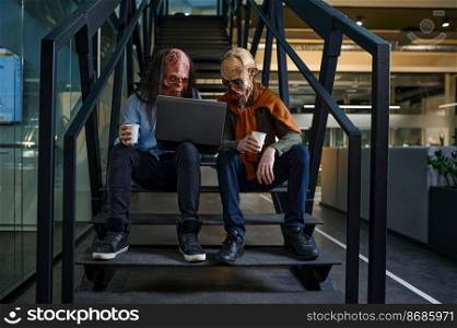 Zombies discussing online project suing laptop and talking sitting on staircase. Short business meeting. Office open space workstation. Zombies discussing online project on staircase