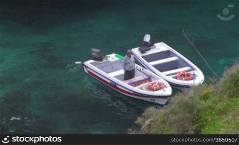 Zwei wei?e Motorboote liegen im flachen tnrkisblauen Meer mit Steinen vor Anker, in einem der Boote steht ein Mann; Knste der Algarve, Portugal.